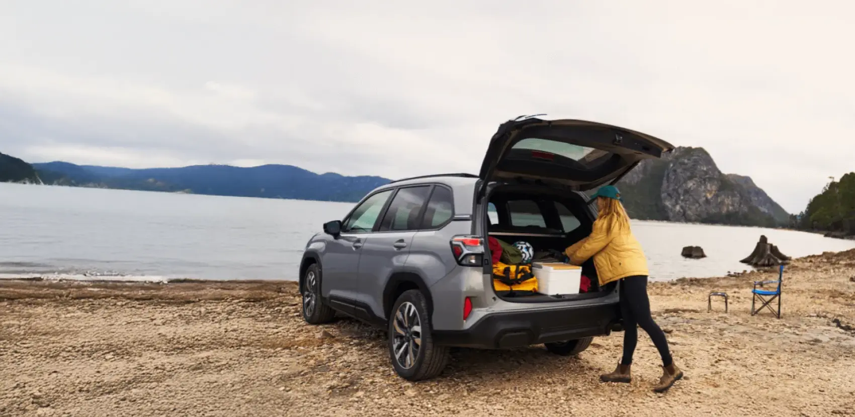 Camioneta Subaru Forester estacionada en frente de lago, con baúl abierto y mujer campista buscando cosas en este.
